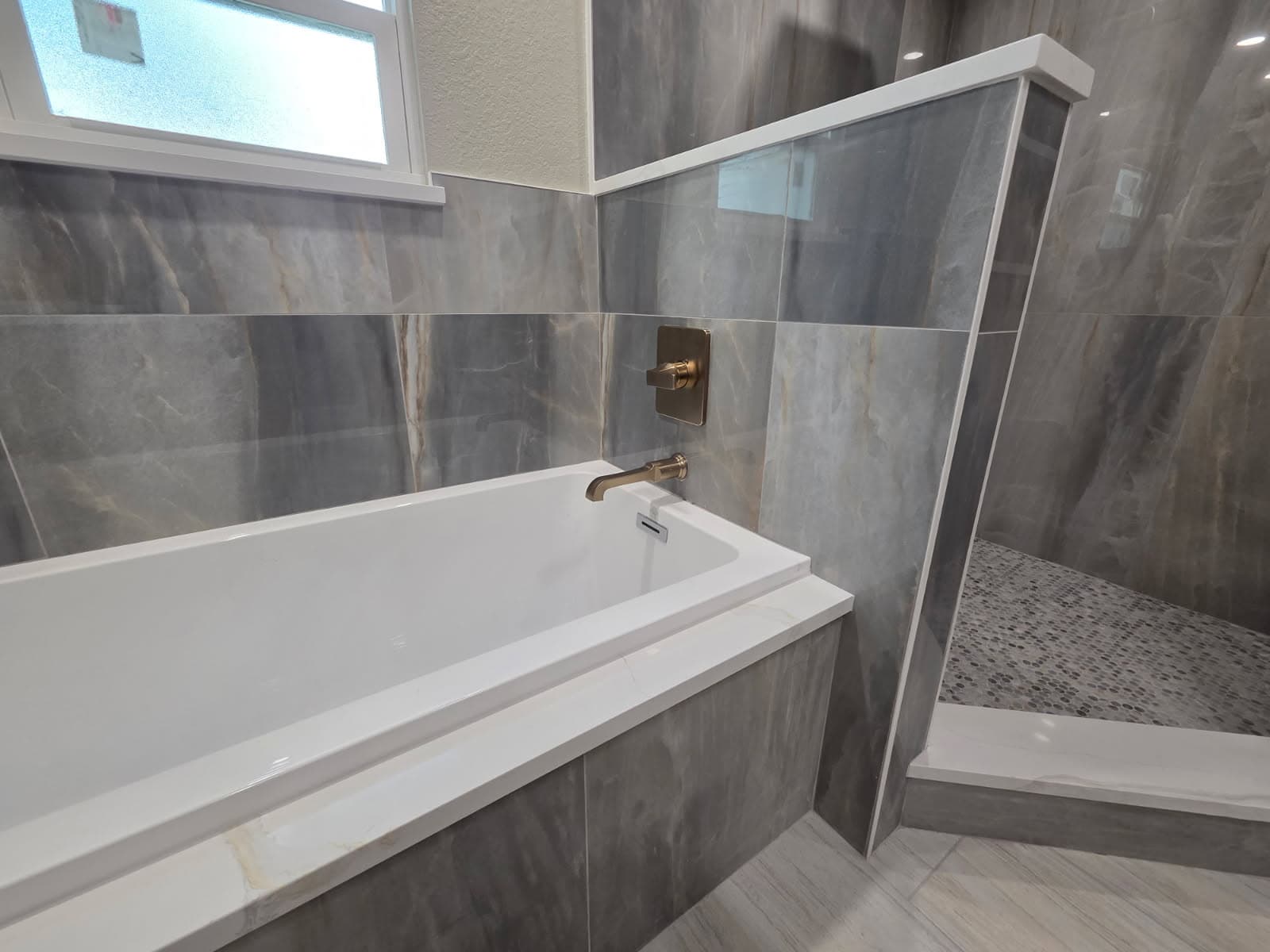 KJB Construction soaking tub with grey marble tile surround and brass wall-mount faucet