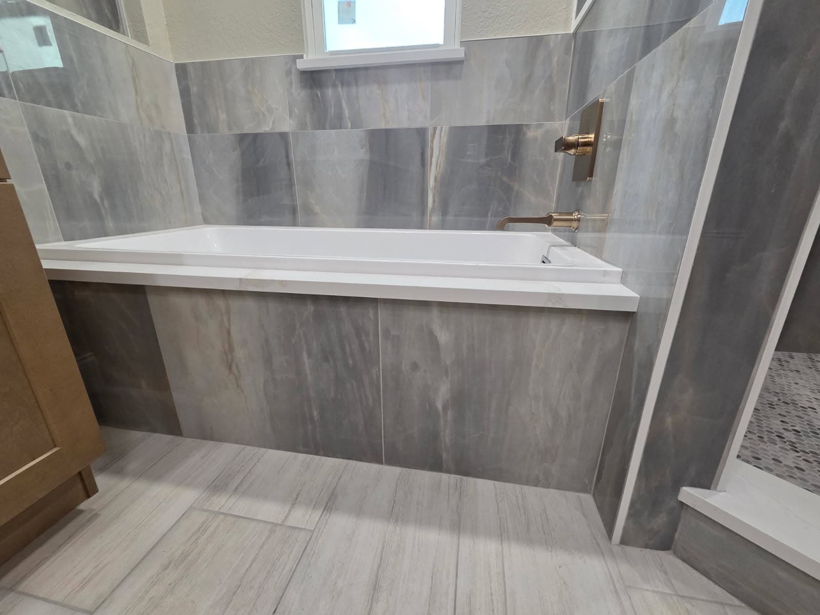 KJB Construction alcove soaking tub with grey natural stone tile surround, brass wall-mount filler, and wood-look tile floor