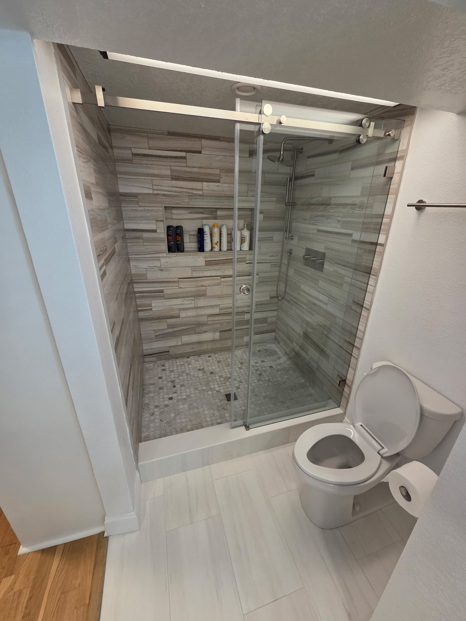 KJB Construction walk-in shower with grey wood-look tile, sliding glass barn door, built-in niche, and hexagonal mosaic floor