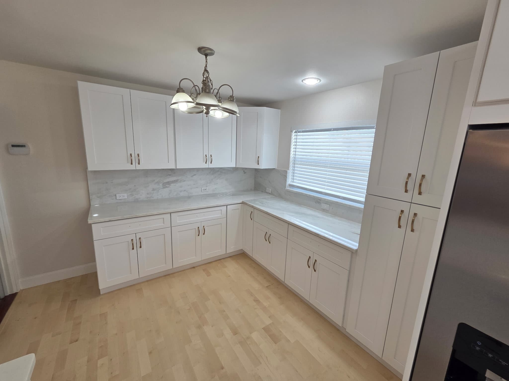 KJB Construction L-shaped kitchen with white shaker cabinets, marble countertop and backsplash, brass hardware, and light hardwood floors