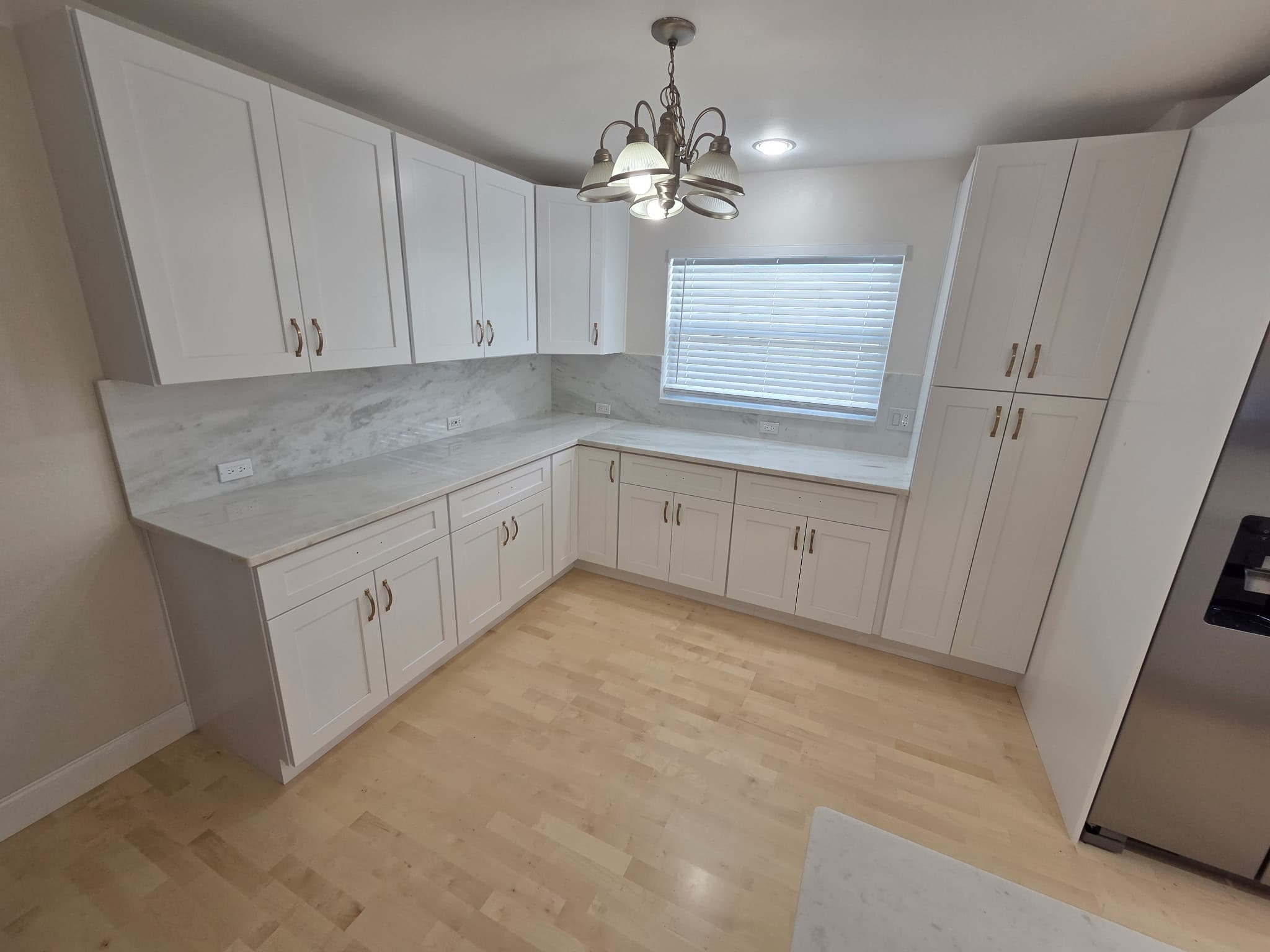 KJB Construction L-shaped kitchen wide view with white shaker cabinets, marble countertop, pantry tower, brass hardware, and stainless appliances