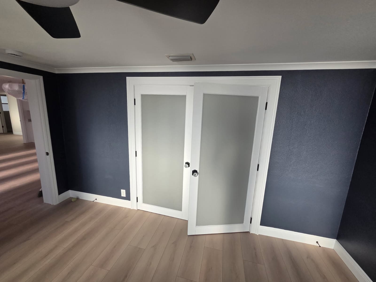 KJB Construction navy blue bedroom with frosted glass French doors, light hardwood floors, and ceiling fan