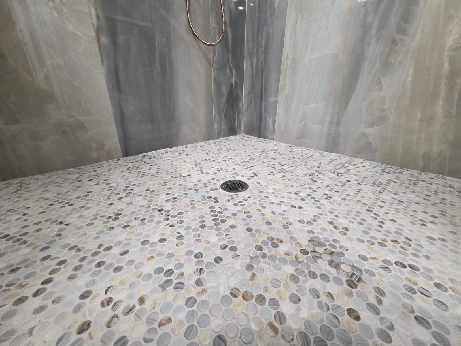 KJB Construction pebble tile shower floor with grey marble walls and brass handheld shower