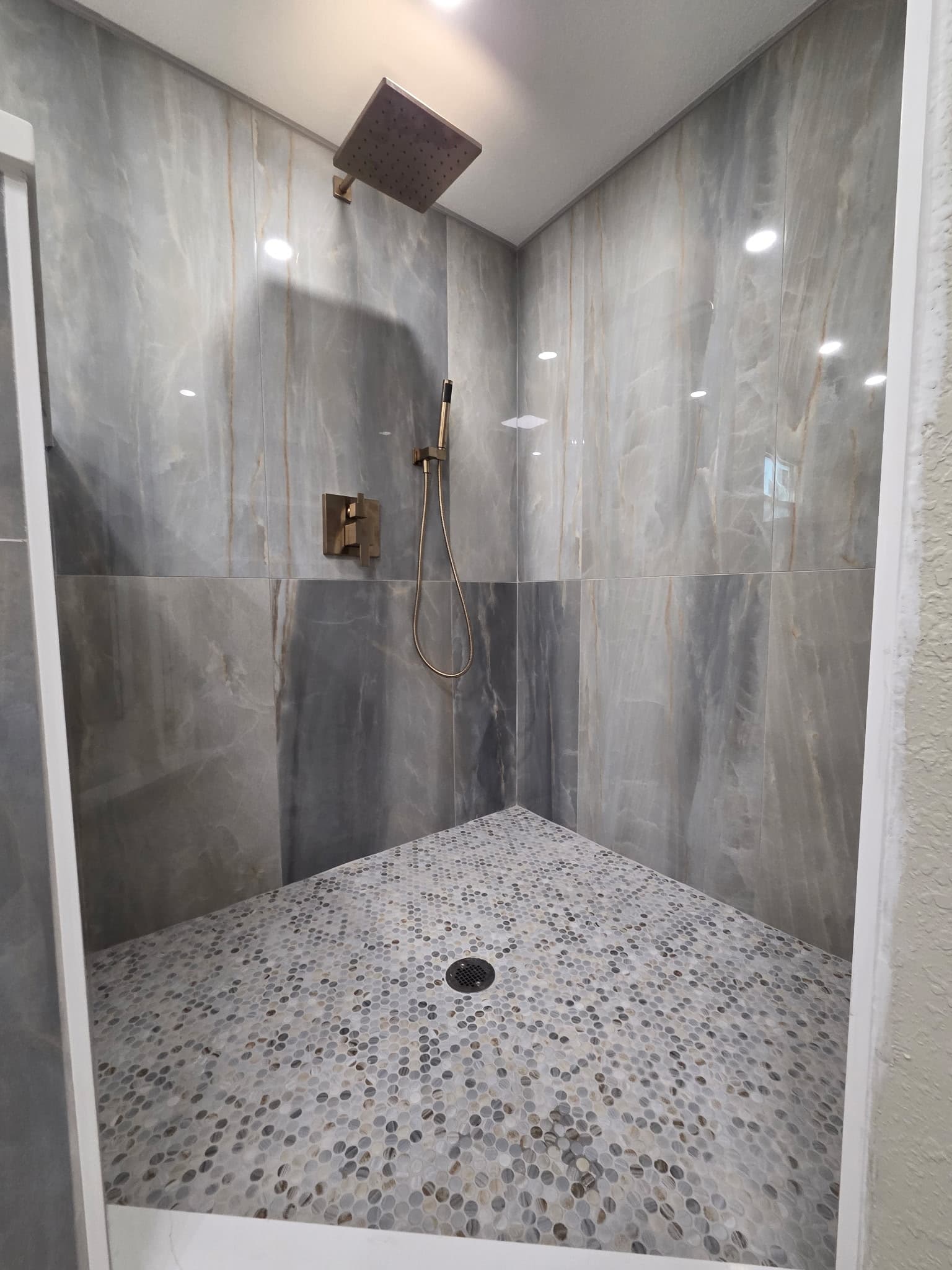 KJB Construction walk-in shower with grey marble walls, pebble floor, and brass rain shower head