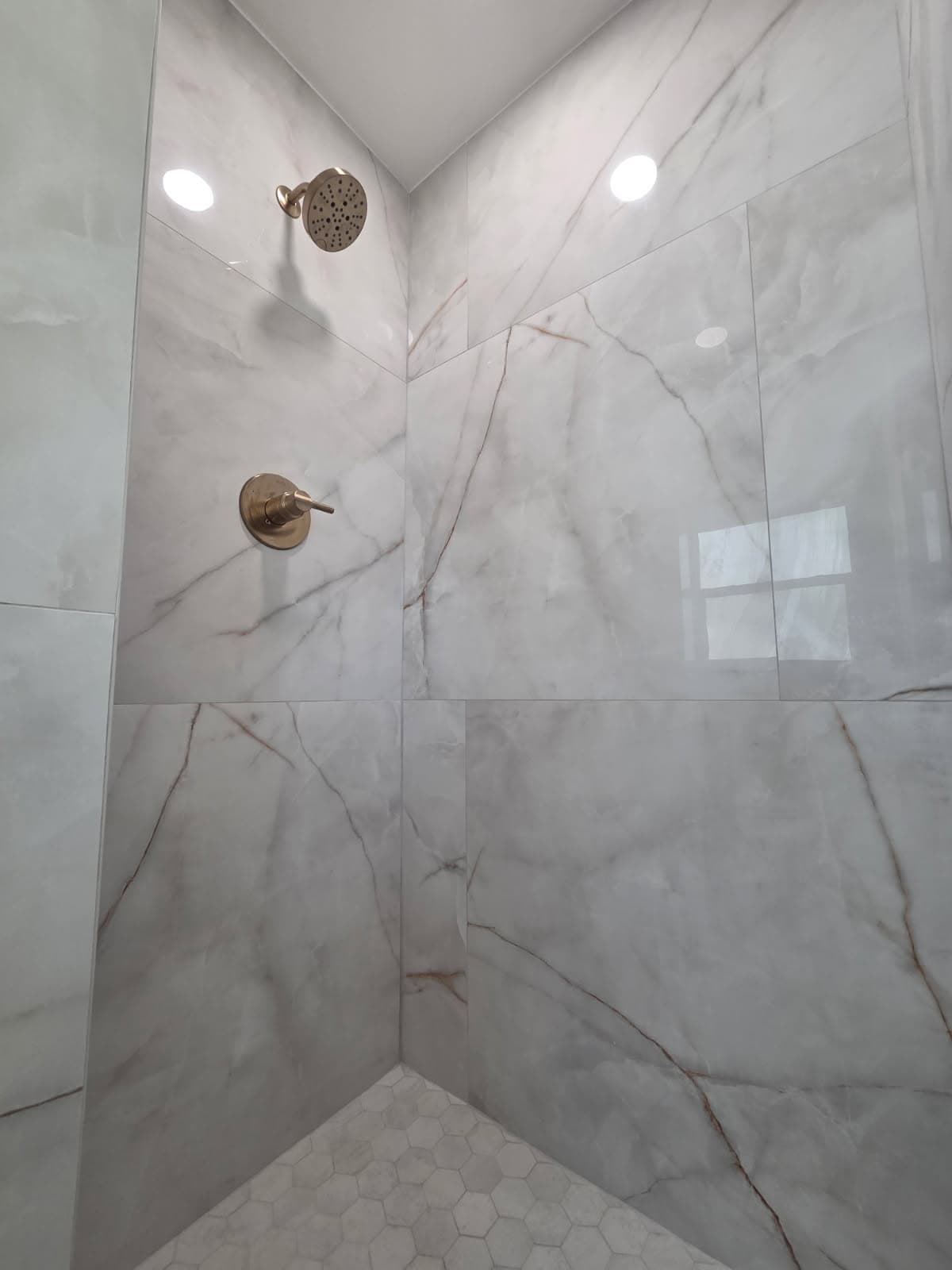 KJB Construction white marble shower corner with brass shower head and valve, hexagonal floor tile, and recessed lighting